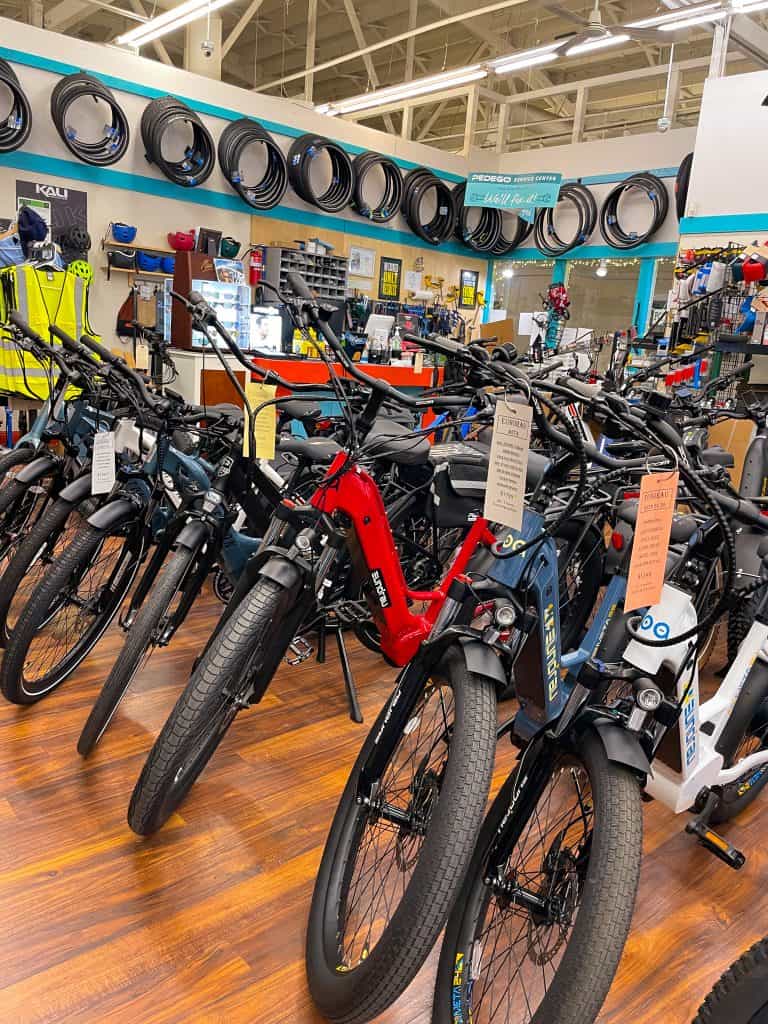 Photo of colorful mountain bikes from the front angle inside All Electric Motion bike shop in Downtown Chico. The background shows various bicycling accessories like reflective vests, helmets, and tire tubes near the checkout counter. The shop has brown hardwood floors and is painted white with teal blue accents.