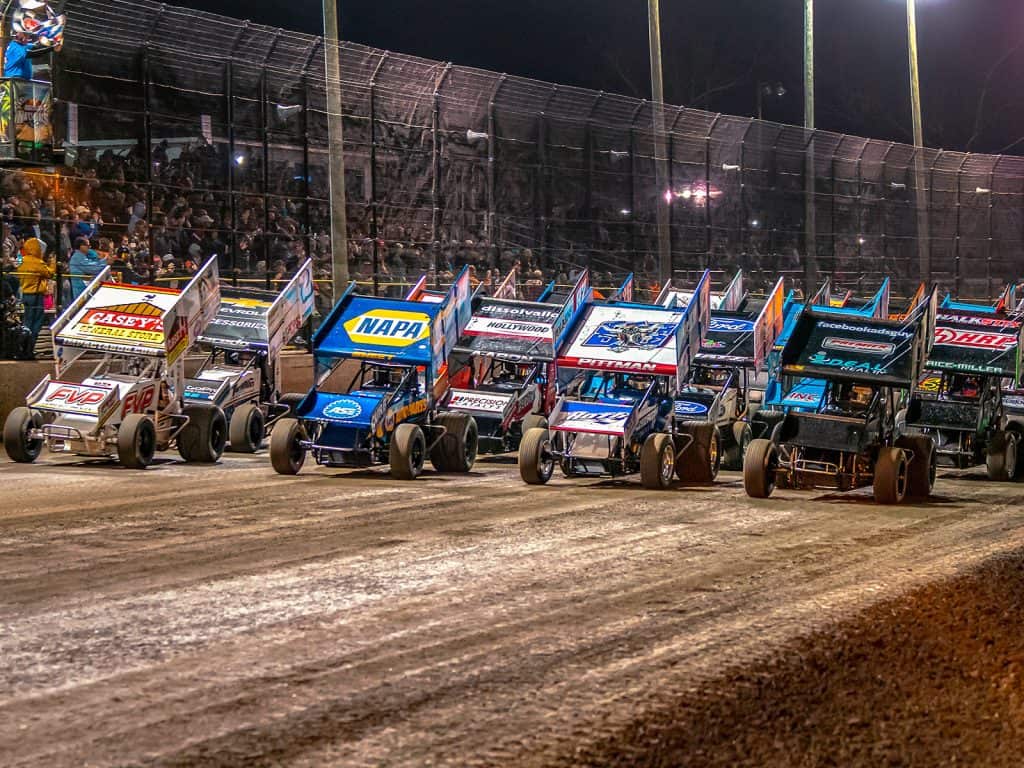 A dozen sprint cars drive pass the flag waver in front of a packed crowd sitting behind high fencing at the Silver Dollar Speedway in Chico, CA.