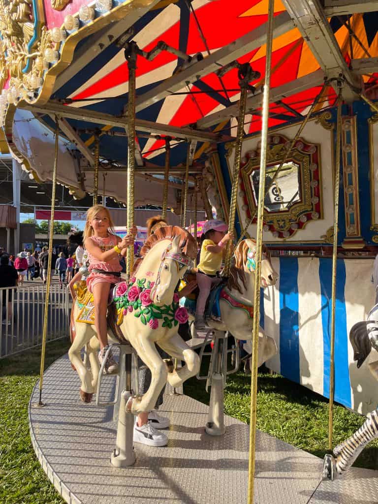 A child smiles while riding the merry-go-round on a sunny summer day with other fair attendees walking in the background at the annual Chico Silver Dollar Fair.