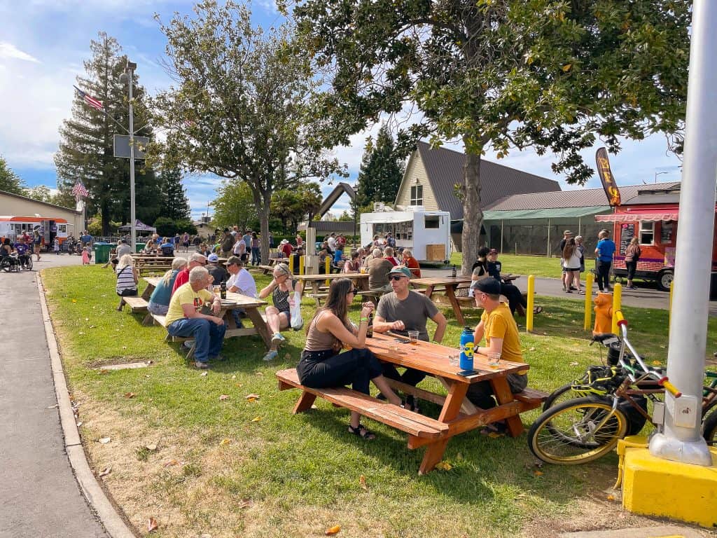 Adults talk on outdoor picnic benches in the sun with bicycles resting in the foreground and food trucks in the background at the Silver Dollar Fairgrounds to celebrate Chico Wildfest.