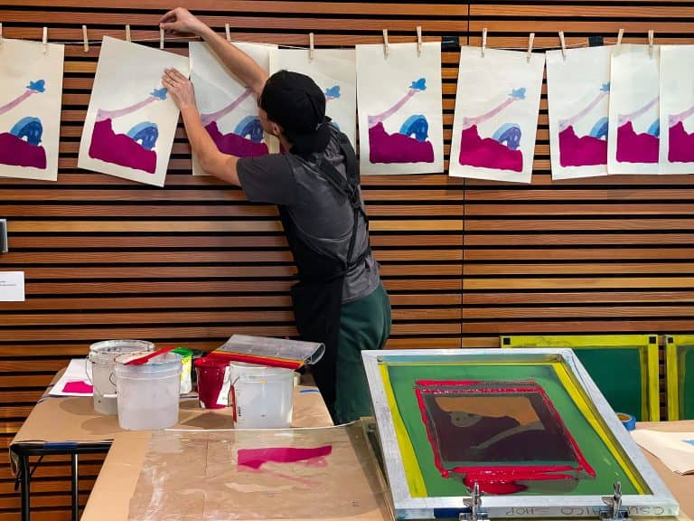 In the foyer of the Janet Turner Print Museum on Chico State University's campus, a young white printmaker wearing a black smock hangs up abstract ink prints on a string with clothespins, with a print machine and colorful supplies in the foreground.