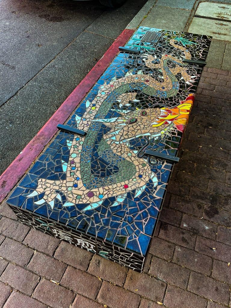 A public bench sits embedded into the sidewalk and shows a Chinese dragon breathing fire made out of mosaic tiles and pieces of mirror. The mosaic background color is black and the dragon is majority tan with small green and purple marble dots and a green and blue belly. The dragon claws and back ridge are mirrors. 