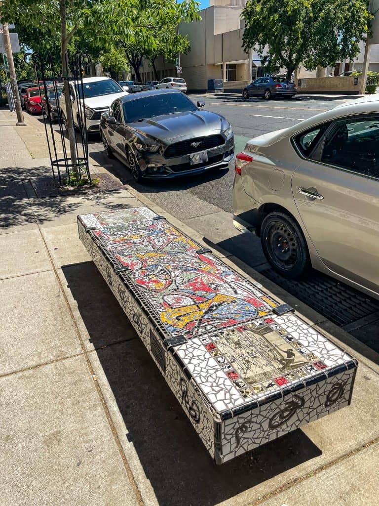 A public bench sits on the sidewalk near a city street lined with parked cars. The bench is covered in mosaic tiles emulating the painting style of Jackson Pollock, with abstract lines in red, yellow, and black on a white background. The bench also includes a tile with Jackson Pollock in the process of painting on the right and leftmost sides. 