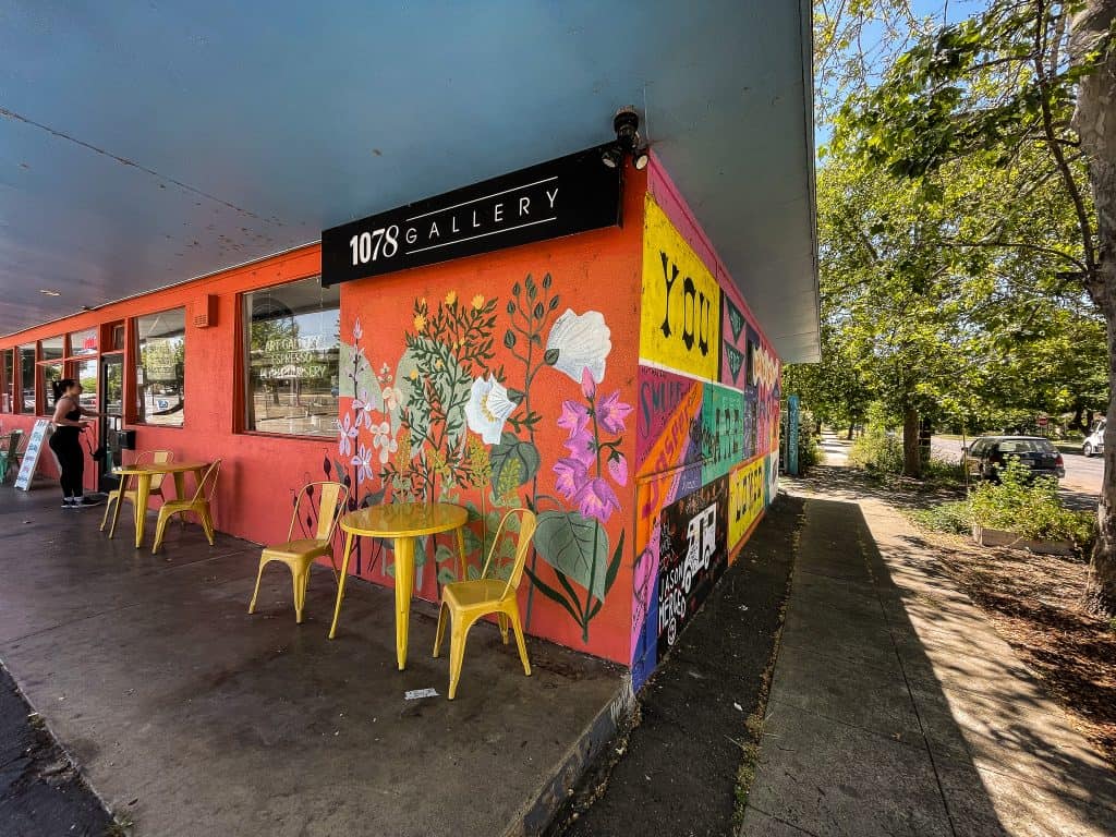 On the right side of the local 1078 art gallery and Equilateral coffee shop, a mural shows native flowers painted onto an orange background and two yellow bistro chairs with a table sit in front of the wall. On the adjacent wall around the corner facing the tree-lined street, is another mural depicting the words “You Are Loved” and bright shapes with names written on them memorializing folks in the community who have passed away.