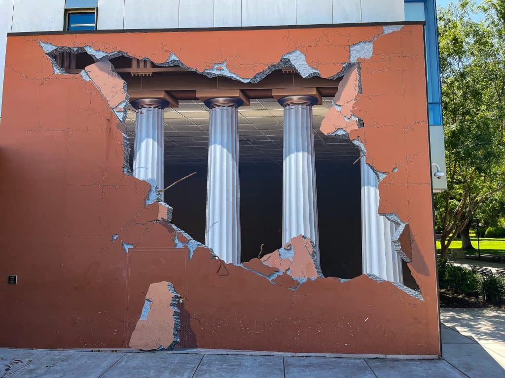 A mural on the side of a Chico State University building shows an optical illusion of a broken concrete wall revealing white Greek pedestals at the entrance of a dark abyss. The mural’s illusion includes a painted on door, iron rebar, and a fallen concrete slab. The painting is completely smooth and the outer wall is painted a terra cotta orange. The mural wall exists near a grassy quad.