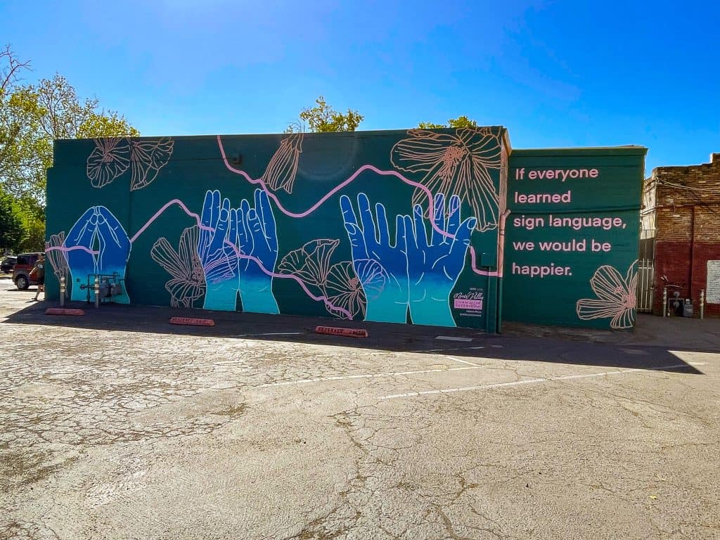 A vibrant mural on the side of a business facade depicts three large hands signing the word “bloom” in American Sign Language. The hands are painted ombre blue and the mural background includes wavy light pink lines and light orange flower outlines. The right side of the mural is cut into the wall and displays the sentence, “If everyone learned sign language, we would be happier” written in pink sans serif font with the teal blue background color.