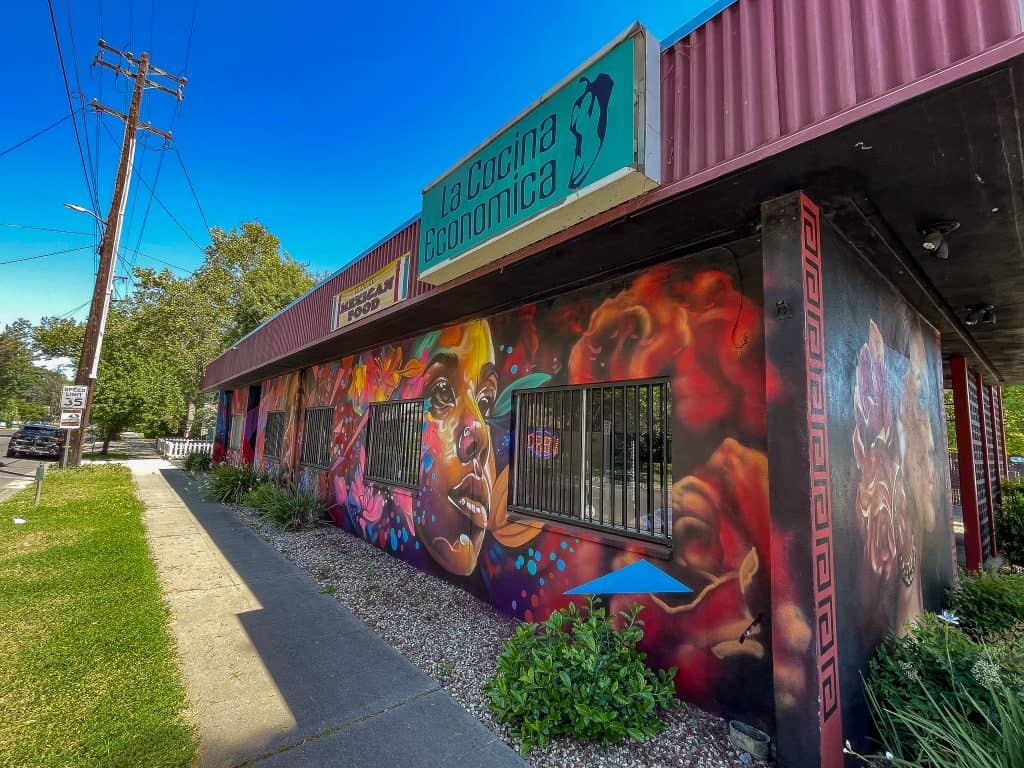 A mural is painted on two sides of a Mexican restaurant called “La Cocina Economica” depicting triangles and dots in bright colors and colorful airbrushed-looking tropical flowers along with two women’s faces looking at a profile view looking forward. The background of the mural is dark red and black abstract flowers that look airbrushed. The building is lined with shrubs and has a sidewalk nearby with a patch of grass lining the street.