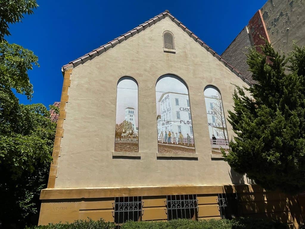 A tryptic mural in arch shapes shows a faded mural of the former Chico Flour Mills industrial building, a handful of men leaning on a nearby wooden fence, and horses and trees in the distance. The mural is painted on the side of the Chico History Museum, where three arch windows used to exist. The museum building is tan with clay tile roofing and shrubs and trees surrounding it.