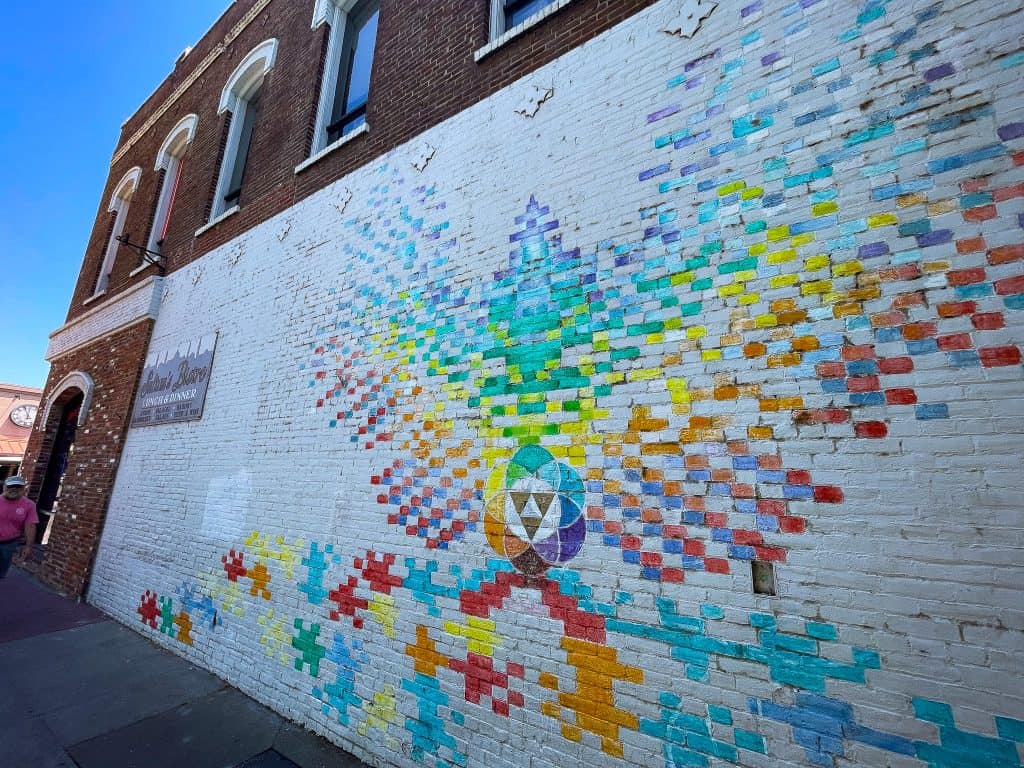 Abstract mural of a phoenix painted in ombre rainbow colors on top of a white painted brick wall. There is a seed of life symbol at the center of the phoenix. The mural is painted on a brick business facade with windows on the top level above the artwork.