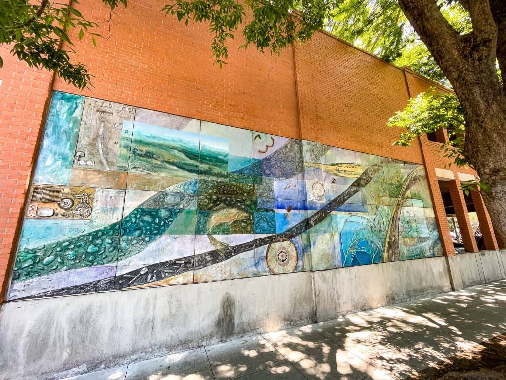 An abstract mural made of fiberglass and painted over in acrylics depicts waterways, green landscapes, and tools. The mural is blocked into rectangles of varying shades of the same earth tone blues, greens, and yellows. The artwork is embedded into a brick parking garage wall and exists on a tree lined street.