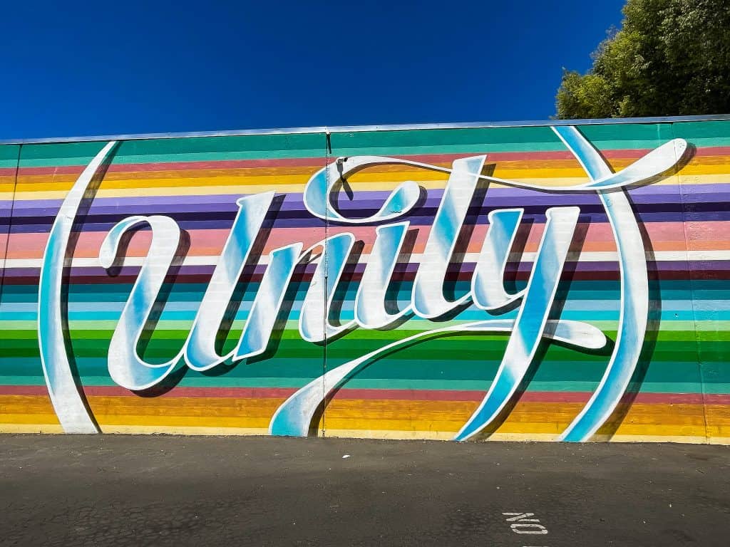 A mural painted on an outdoor wall facing a parking lot depicts the word “Unity” in cursive font with a background of horizontal stripes in various rainbow colors out of sequence. The word “Unity” is painted to look metallic and shiny with a shadow as if the word is raised from the wall surface.