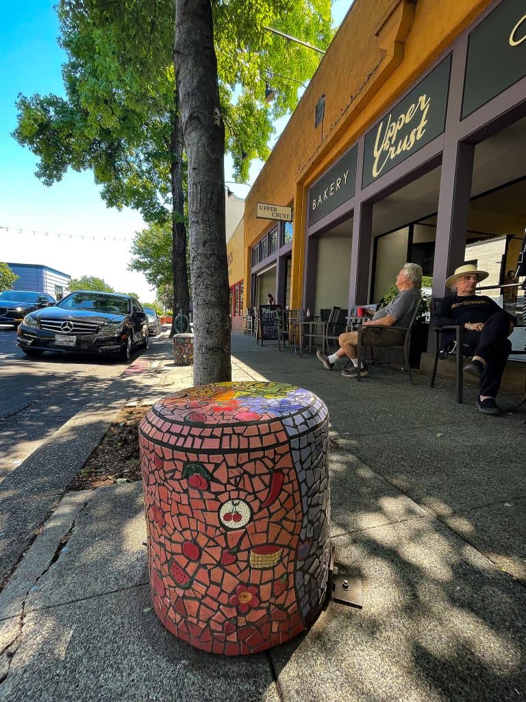 A mosaic pedestal sits on the sidewalk of a city street, in front of a local coffee shop where patrons are eating outside on bistro tables. The pedestal is covered in colorful tiles showing different fruit and vegetables like cherries and peppers, all organized by the colors red, purple, green, and orange. The Music Pedestal is visible in the background. 