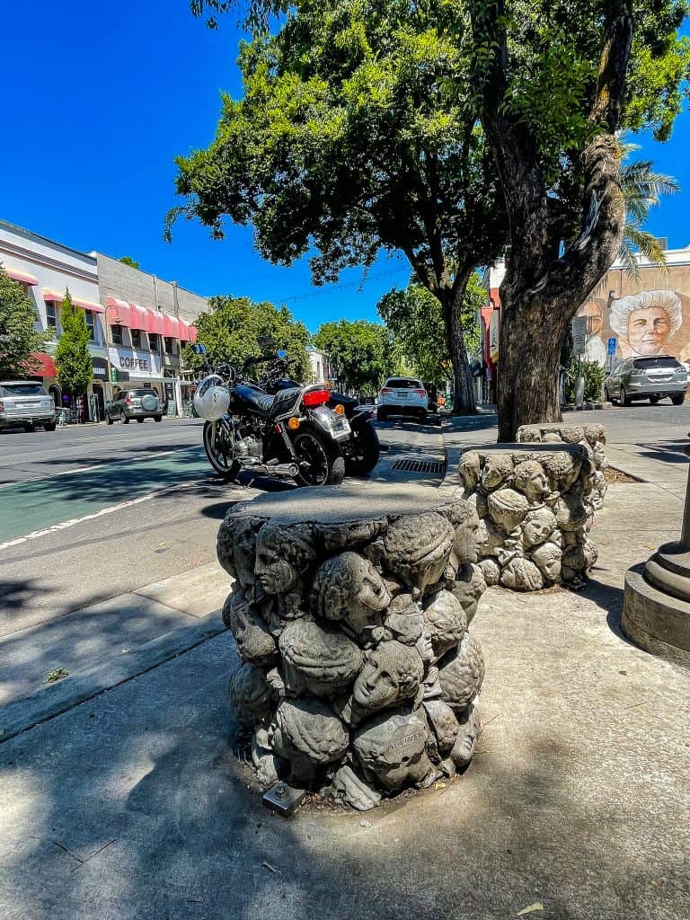 Three staggered pedestals sit on the sidewalk next to a city street where cars and motorcycles are parked in the distance. The pedestals are made of concrete and show 3-D Greek statue style heads stacked on top of each other, all the size of softballs. The top of the pedestal is smooth and flat to sit and the sides are bumpy with the heads contours. 