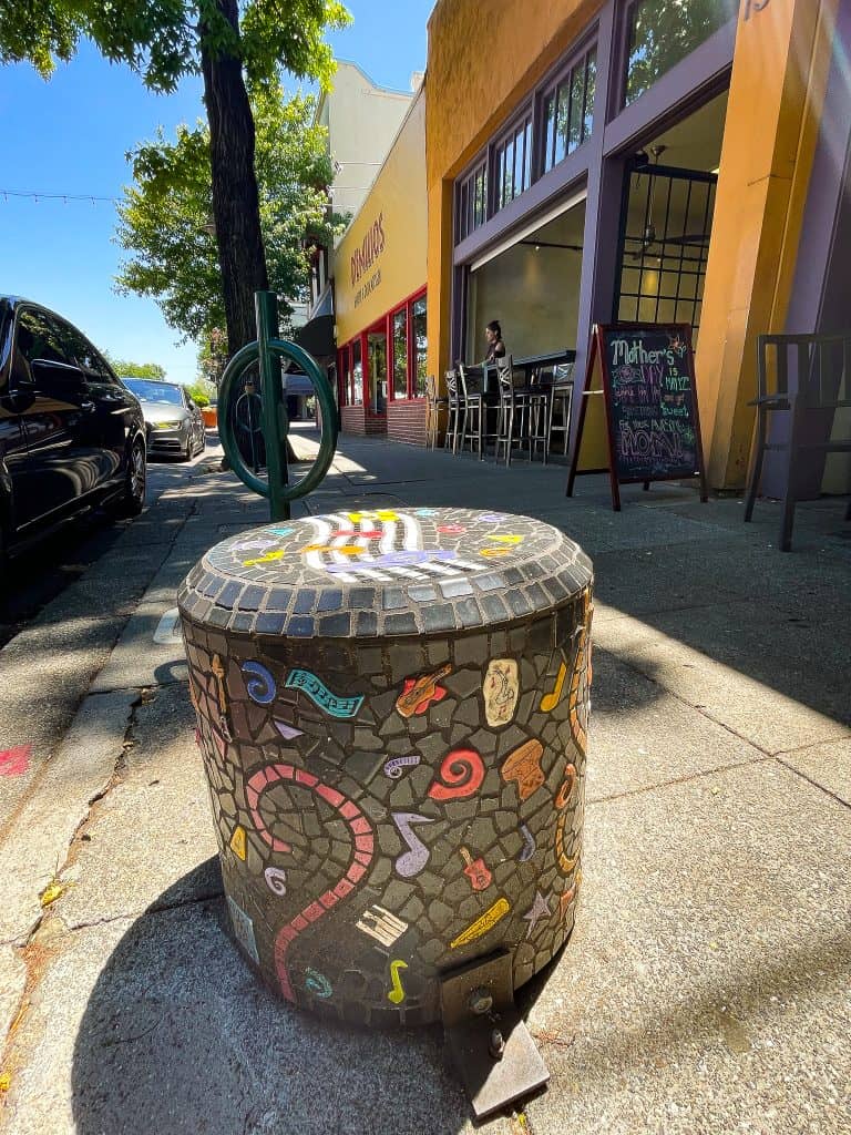 A mosaic pedestal sits on the sidewalk of a city street, in front of a local coffee shop. The pedestal is covered in mosaic tiles depicting various musical symbols like music notes, guitars, and sheet music, all in bright colors on a black background. A few abstract spirals fill in the art space. The symbols are mainly yellow, red, white, and purple.