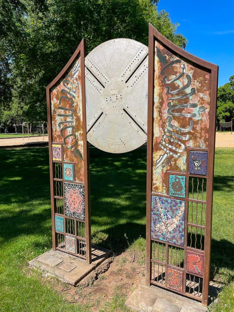 An abstract, slightly rusted metal gate with two rectangular side panels containing colorful mosaic tiles depicting cultural symbols and a large circle that looks like a sanddollar at the center joining the two panels sits in a grassy park field under the shade of nearby green trees on a sunny day.