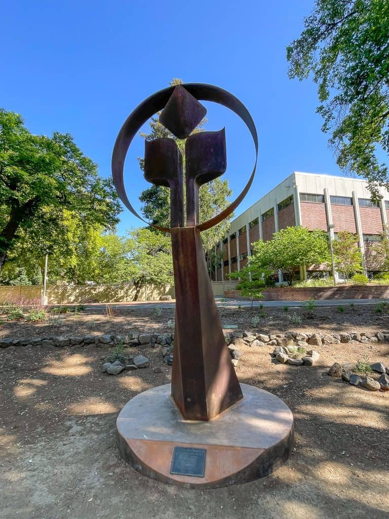 A tall steel sculpture sits in a rock garden with red brick buildings and green trees in the background. The sculpture depicts an abstract torch and flame.