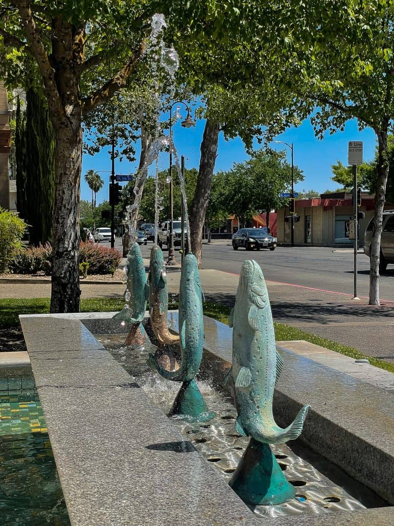 Oxidized green salmon sculptures spout water into a long rectangular fountain made of grey granite and colorful green, blue, and yellow tiles installed next to a busy city tree-lined street pictured on a sunny day.