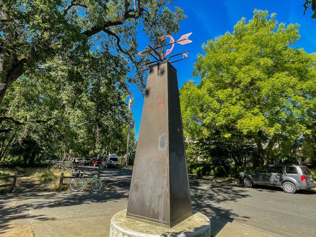 A tall metal sculpture stands at a sidewalk corner bordering a green park and residential houses. The sculpture works as a bike rider’s guide, depicting common local destinations and how many miles away they are. It has a bike gear shaped compass at the top.