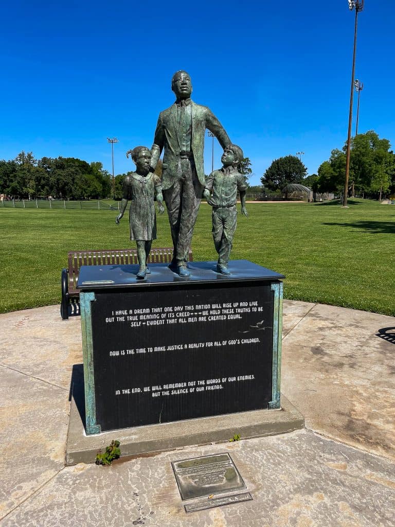 Large bronze sculpture of Dr. Martin Luther King Jr. walking with two children standing at his sides and his hands on their backs as if guiding them forward sits atop a black pedestal with Dr. Martin Luther King Jr.’s “I Have a Dream” speech engraved on the front. The sculpture has benches surrounding it and stands in the middle of a green park field.