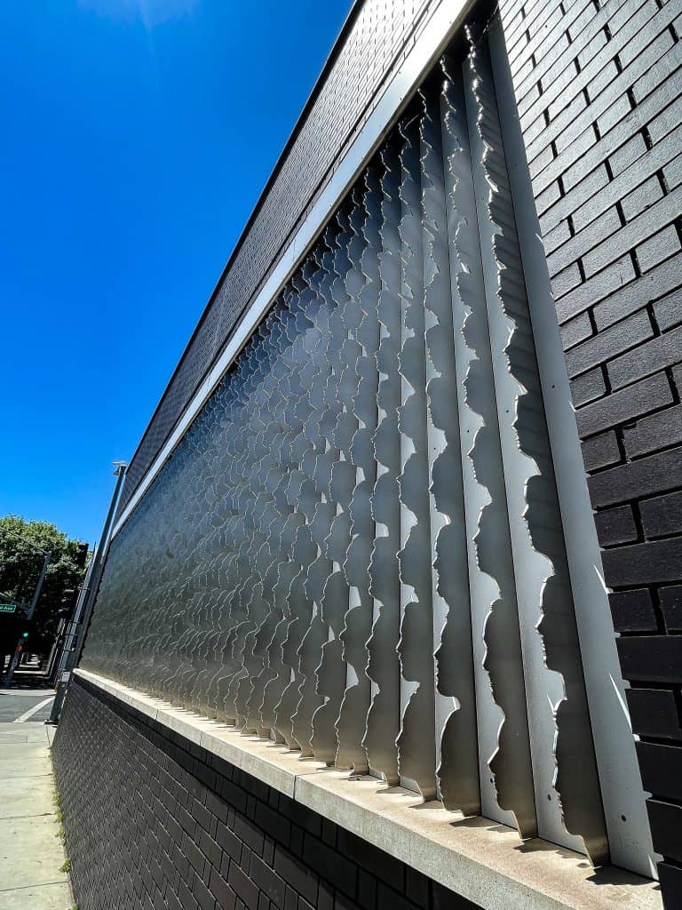 Large sculpture of layered human profiles made of metal, visible only at a close vertical angle. The sculpture is embedded into a black brick wall on an outdoor facade adjacent to a city street, pictured on a sunny day.