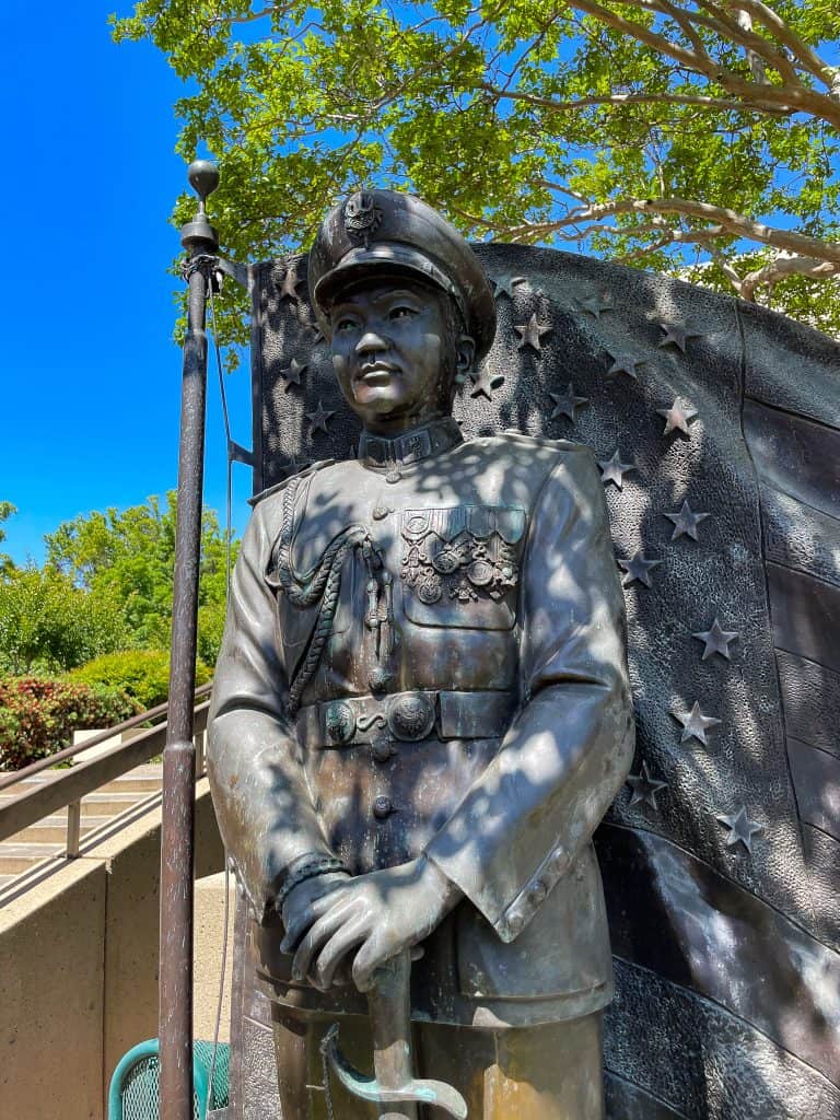 Large bronze full-body sculpture of General Vang Pao in full military uniform with a bronze waving American flay in the background pictured on a sunny day.