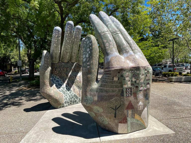 A giant marble sculpture of two open hands facing each other posed like a round ball exists between the hands. The sculpture is embedded into the sidewalk and the hands are designed with scenes of abstract waterways, trees, and houses embedded into the stone with light colored terrazzo tiles. Pictured on a sunny day in front of the City of Chico Municipal Building parking lot.