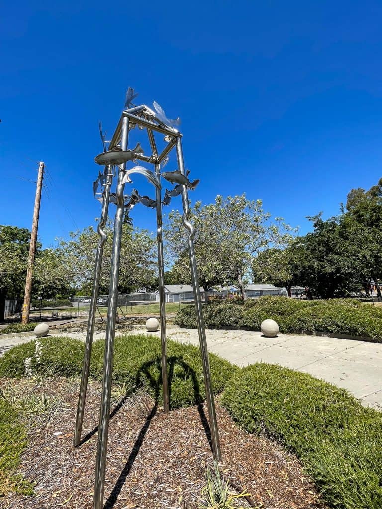 Tall shiny silver metal sculpture made of four posts joined at the top in a tetrahedron shape with silhouettes of salmon attached to the top third of the sculpture. The piece stands in a public garden with green grassy shrubs and concrete spheres embedded into the sidewalk.