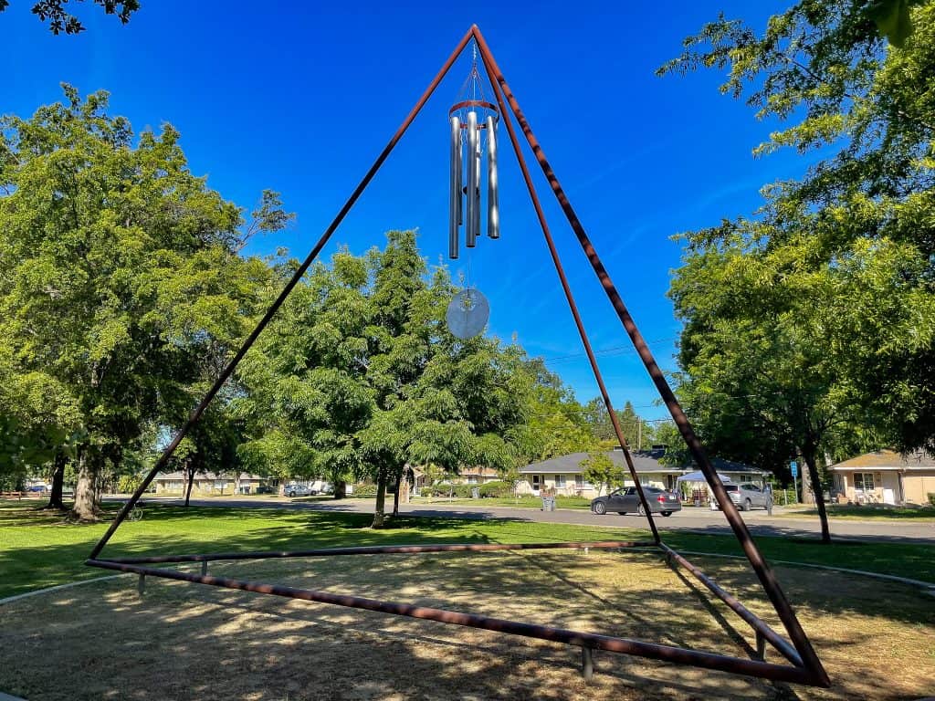 A giant metal tetrahedron shape stands in the middle of a grassy field with many nearby green trees near residential homes. In the top point of the tetrahedron is a large metal windchime with a large abstract circle hanging down.