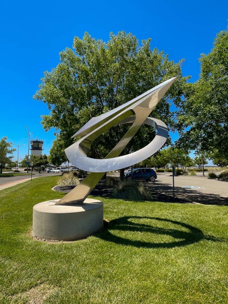 In front of the Chico Regional Airport, in a grassy area, a shiny silver sculpture stands depicting an abstract plane shaped like a paper airplane flying through a circle.