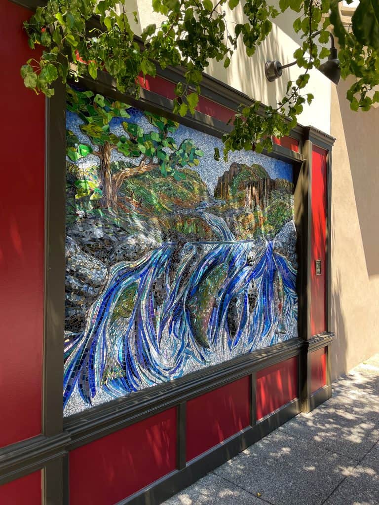 Chico Public Art: Colorful 3-D glass mosaic depicting a salmon-filled creek running from green mountains and lined with trees installed on an outdoor business facade with leaves from a nearby tree in the foreground.