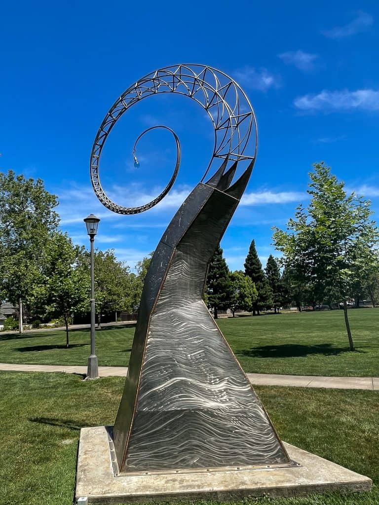 Sparkly silver spiral sculpture sits in a green park field with trees in the background. The structure is covered in metal at the base and is exposed frame skeleton at the top third. A small lightbulb ornament hangs from the tip of the spiral.