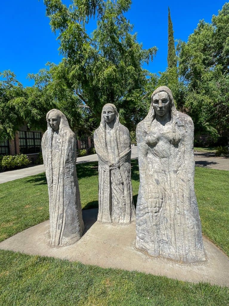 Three staggered concrete sculptures depict three women in long garments and long hair with slightly downcast eyes installed in a grassy quad on Chico State University campus among green deciduous trees.