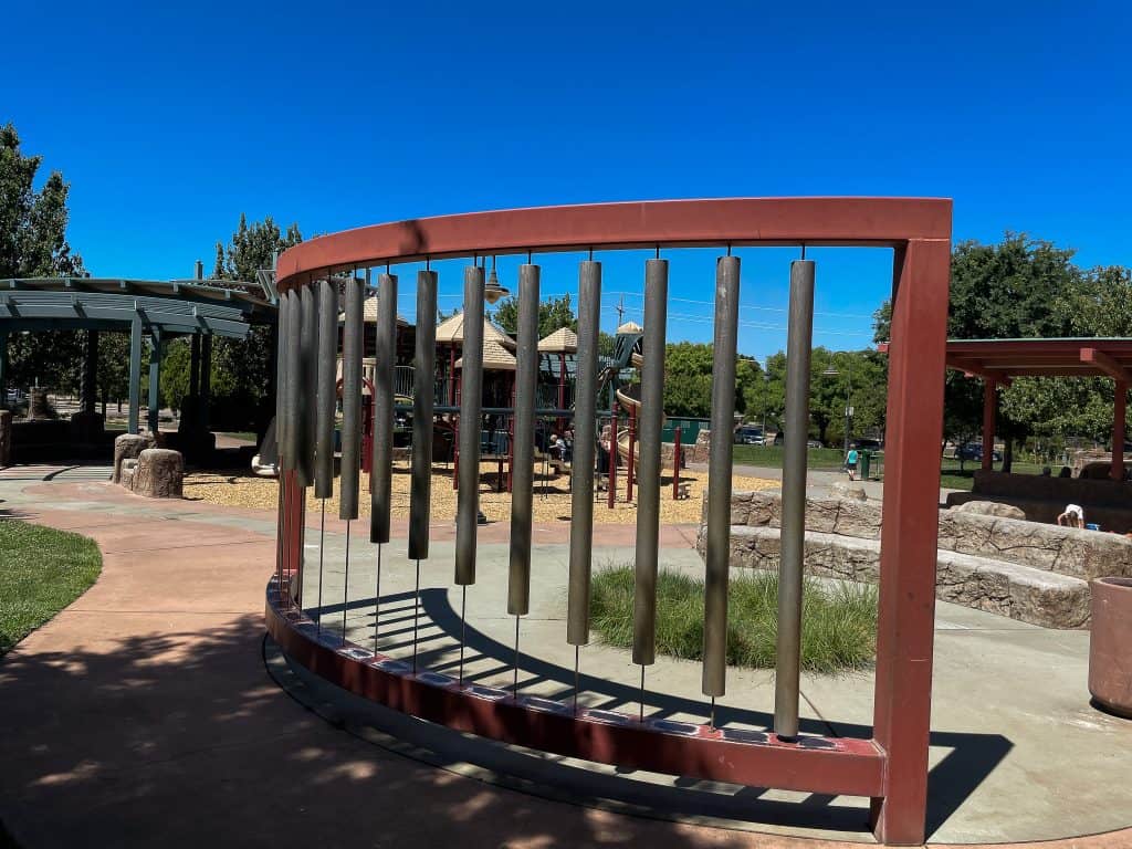 A giant metal xylophone with a curved red frame stands in the middle of a public park shaded by adjacent trees, with a playground in the background on a sunny day.