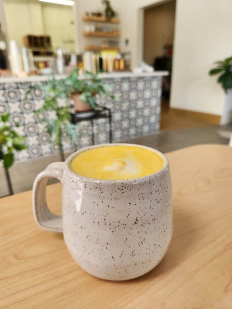 Turmeric chai latte in a speckled ceramic mug sits on a wooden table at the Chico Chai chaihouse. The background shows a cafe counter with indoor plants.