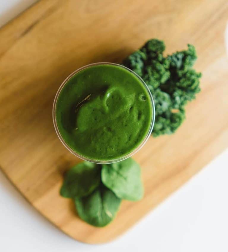 Aerial photo of a green smoothie in a tall glass sitting on a brown cutting board with leaves of kale and spinach resting next to it at Fresh Twisted Cafe