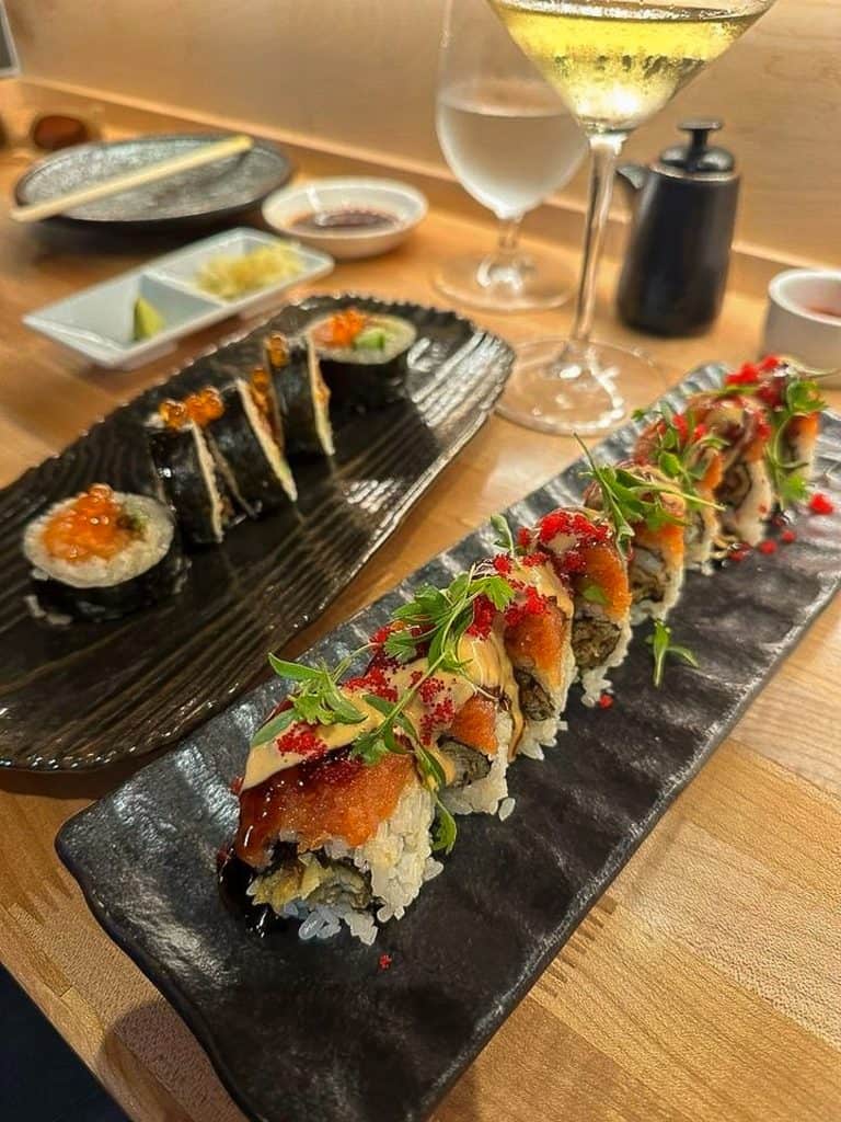 The image shows two sushi rolls on black plates with a white wine glass, water glass, and side dishes in the background at Nama Sake Sushi & Restaurant. The roll in the foreground has rice on the outside and is topped with roe, a spicy mayo sauce and sprigs of cilantro. The roll to the left of the image has roe on the inside and is wrapped with nori seaweed on the outside.