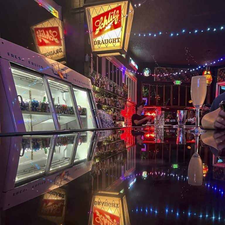 Wide-lens image of the Winchester Goose bar at night. The bar counter is shiny vinyl and lit by string lights that hang above. Behind the bar is a vintage grocery cooler now filled with beverages to order. In the foreground sits a champagne flute and in the background a bartender wearing a hat chats with two patrons.