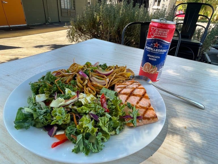 Image of a dish including a grilled salmon filet, noodles, and kale salad with a canned grapefruit sparkling beverage to the right of the white plate at Bacio Chico. The plate sits on a light wood table outside with metal bistro chairs and shrubs in the background.