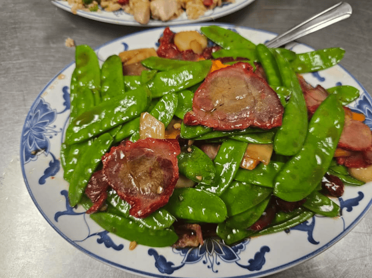 Image of family style dish of sauteed snap peas and roasted pork on a blue and white plate sitting atop a stainless steel surface with another dish blurry in the background from Happy Garden Restaurant.